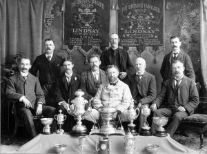 Neuf hommes assis devant des trophées et bannières de championnat.