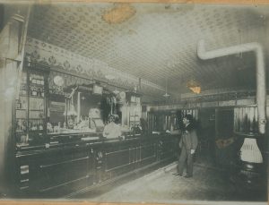 Un barman portant un manteau blanc derrière un bar d’hôtel sert un client qui porte un complet noir et un chapeau.