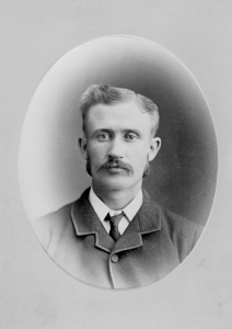 Photographie de studio d’un jeune homme, cheveux et moustache peignés, qui porte un complet noir.