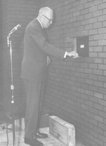 Un homme portant des lunettes, debout derrière un microphone, sur le point de poser une brique dans le mur d’un bâtiment.