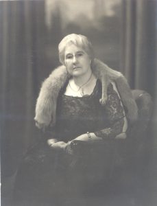 Une femme grisâtre porte des lunettes, une robe noire à dentelle et une fourrure de renard autour du cou, assise sur une chaise.