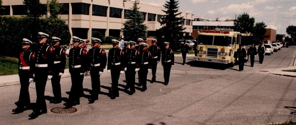 De nombreux membres de la garde d’honneur marchent en uniforme de grande tenue, devant un camion d’incendie jaune, en tête du cortège funèbre de Morley James. 
