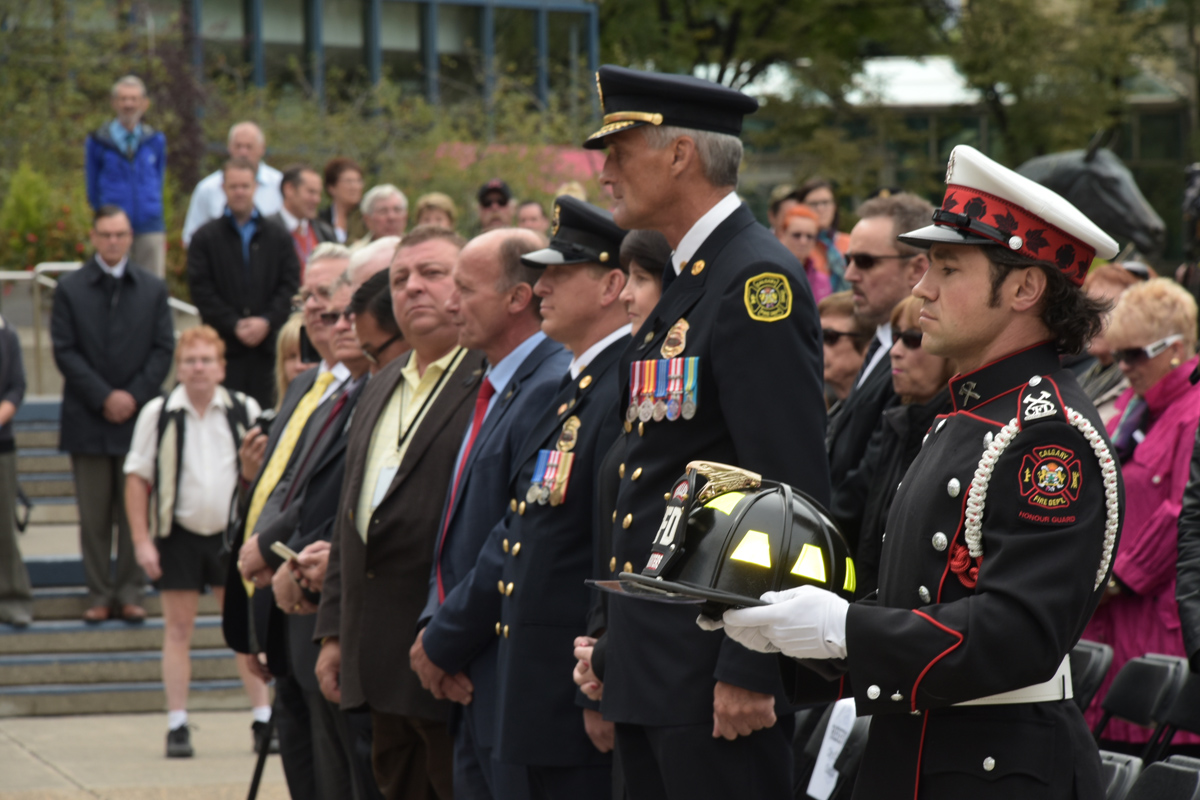 John Judge, garde d’honneur en uniforme de grande tenue et gants blancs, tient le casque commémoratif orné d’insignes jaunes à la place de l’hommage (Tribute Plaza). En arrière-plan, on aperçoit le chef du service d’incendie et la foule.