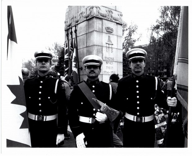 Trois gardes d’honneur portent des drapeaux et une hache cérémoniale devant le monument de la Première Guerre mondiale au parc Central Memorial. Cette photo est en noir et blanc.
