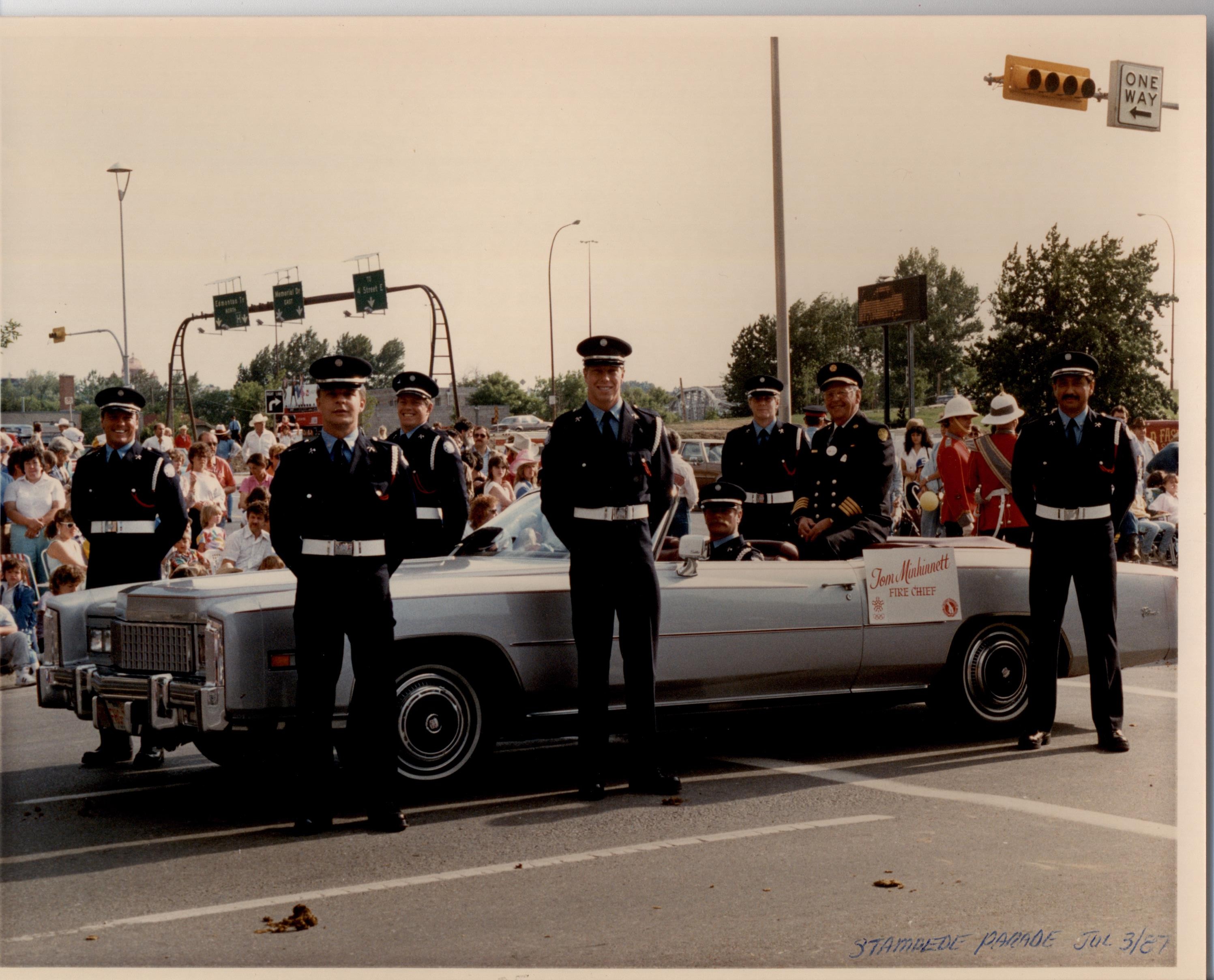 Six membres de la garde d’honneur, debout à côté d’une voiture décapotable dans laquelle le chef des pompiers est assis. On aperçoit des spectateurs en arrière-plan.