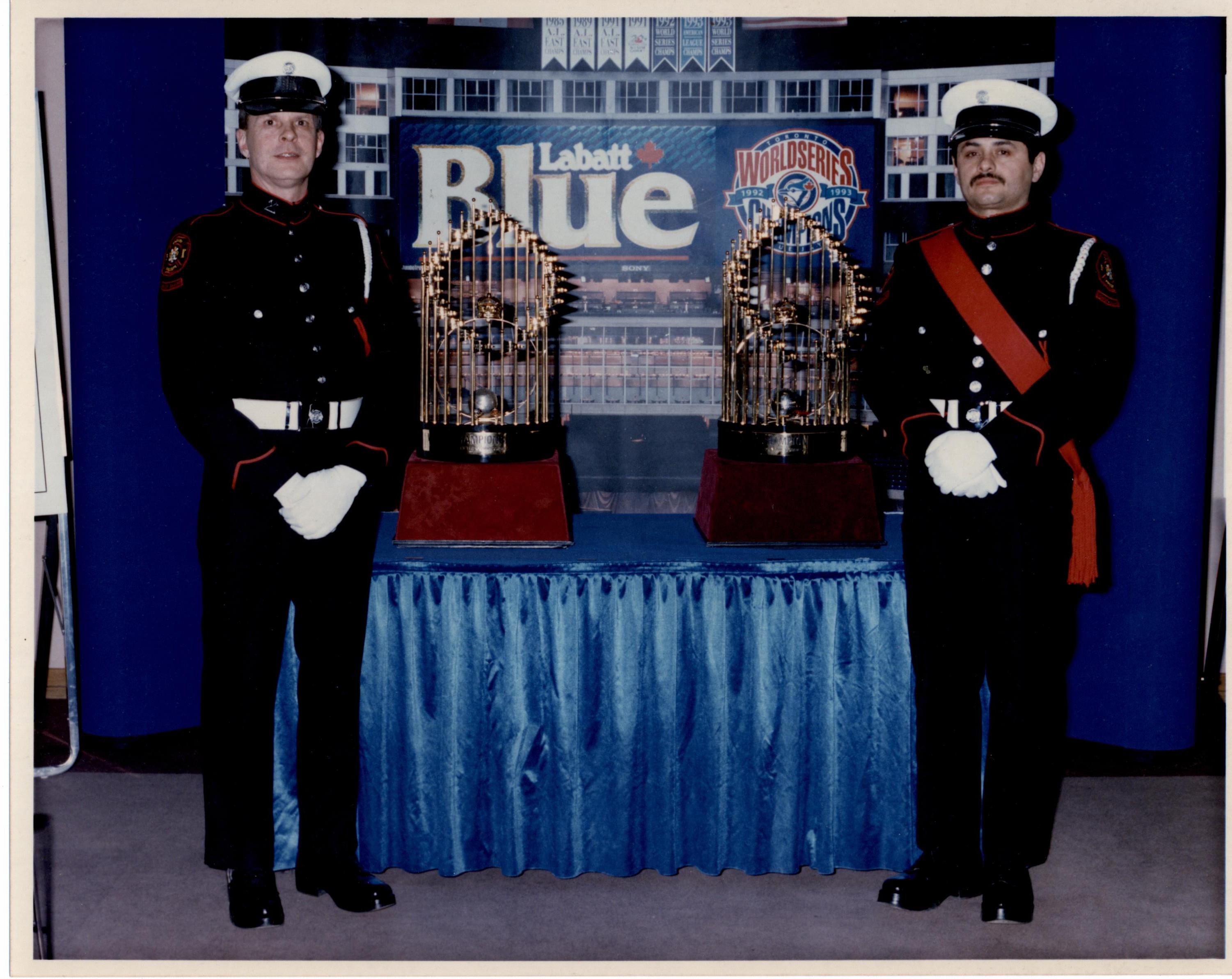 Rick Choppe et Shannon Pennington, membres de la garde d’honneur, avec les trophées de la Série mondiale, 1993, tous deux en uniforme de grande tenue Les trophées sont placés sur une table drapée de bleu, avec arrière-plan bleu.