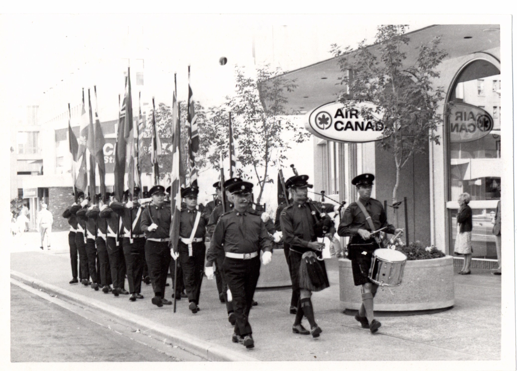 Honour Guard parades past palliser square in black and white photo, most bearing flags, and led by pipes and drums.