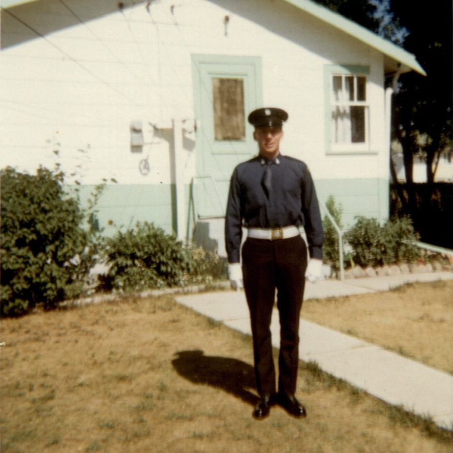 Dennis McIvor, sur le parterre d’une maison blanche et bleue portant l’uniforme original numéro un de la garde d’honneur, avec cravate rentrée dans sa chemise