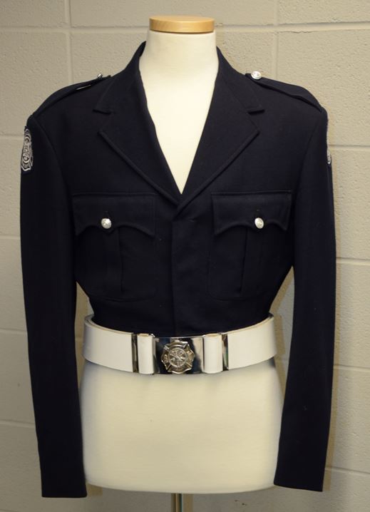 Uniforme original de grande tenue numéro un de la garde d’honneur  Veste noire de style combat munie de l’insigne du service d’incendie de Calgary sur chacune des épaules, de deux pochettes à la hauteur de la poitrine, de plis creux, de rabats festonnés et de deux épaulettes fixées à l’aide de boutons argentés Ceinture en cuir blanche et boucle plaquée argent sur la veste portée par un mannequin blanc sans tête