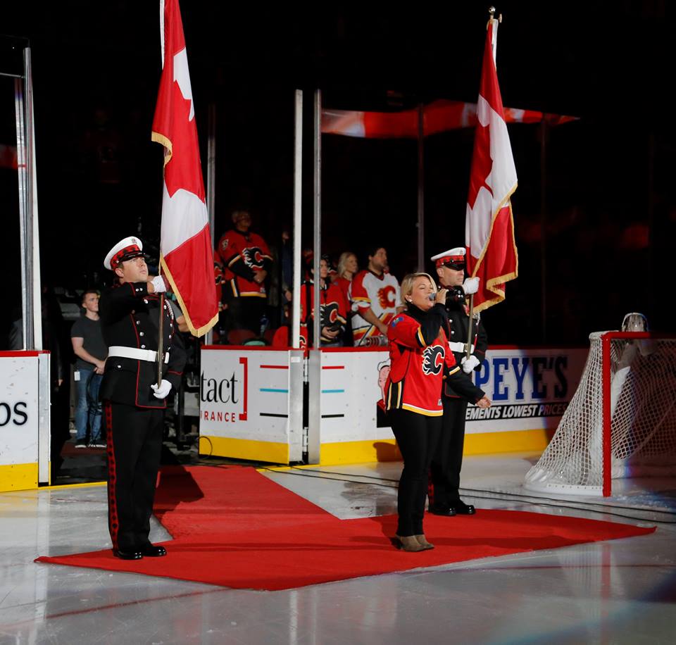 Deux membres de la Garde d'honneur se tiennent à l'attention en tenue complète chacun portant le drapeau canadien tandis que la chanteuse blonde chante l'hymne national dans un chandail des Flames de Calgary.