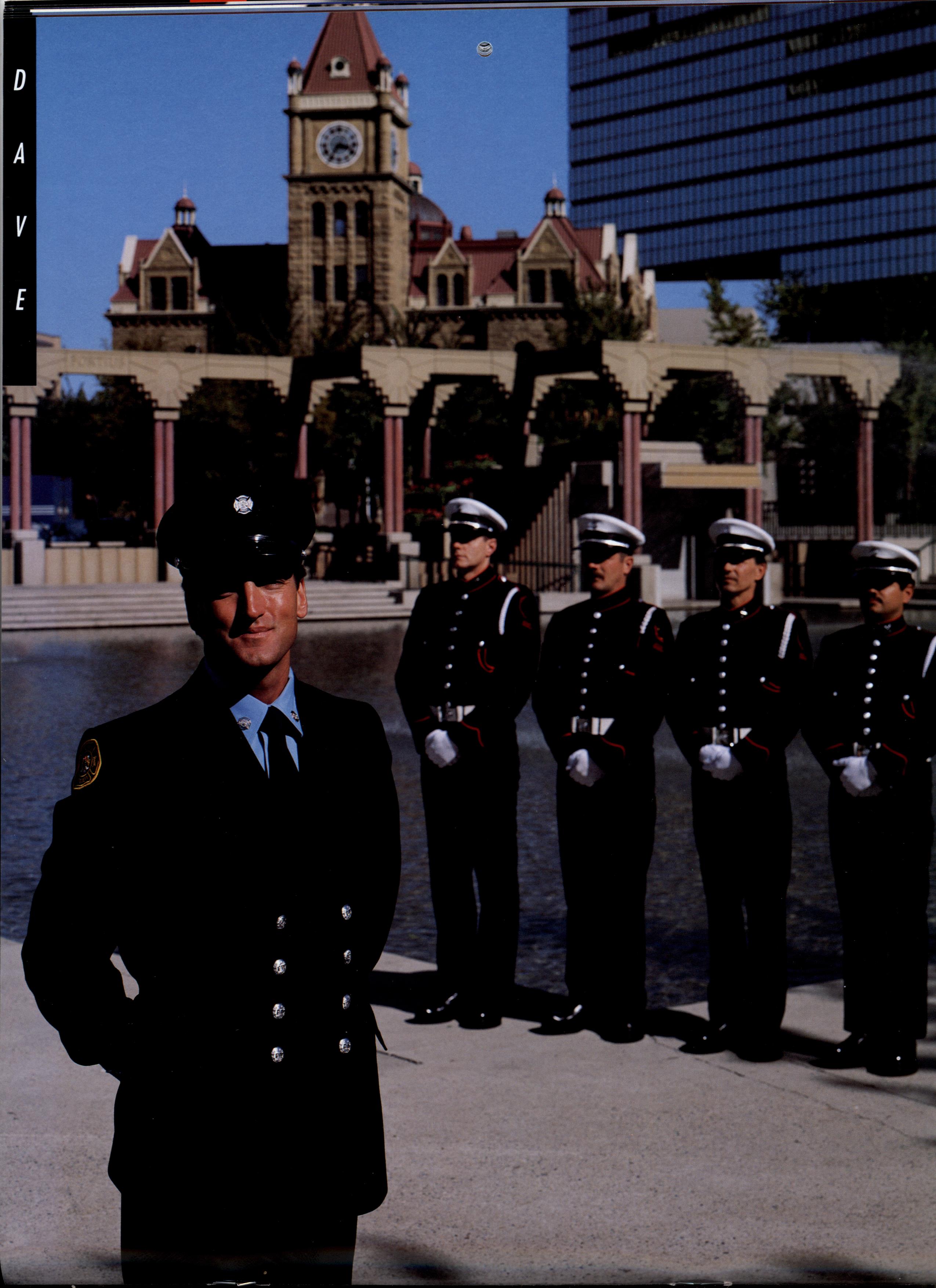 Un pompier debout à gauche, en avant-plan, et quatre membres de la garde d’honneur se tiennent au garde-à-vous un peu plus loin. En arrière-plan, on aperçoit la place olympique et l’hôtel de ville historique en grès.