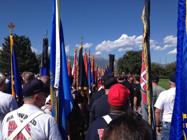 Des gardes d’honneur en tenue décontractée se préparent à participer au cortège commémoratif (le dos à l’appareil photo), drapeaux à la main. On aperçoit les montagnes en arrière-plan.