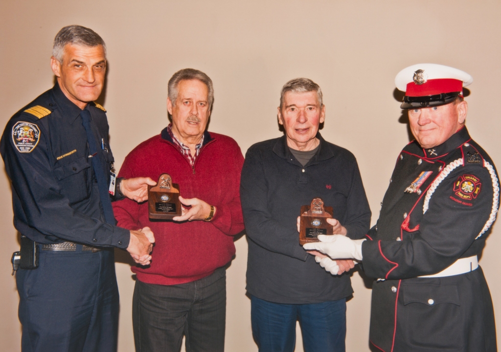 Les fondateurs Brian Freney et Dennis Mcivor, vêtus de tenues décontractées, reçoivent leurs plaques commémoratives par deux pompiers en uniforme.