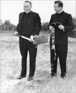 Photo noir et blanc de deux prêtres à une cérémonie d’inauguration, l’un avec une pelle, l’autre avec un livre.