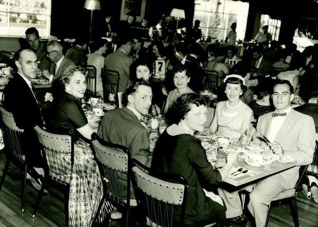 Photo noir et blanc d’un groupe d’hommes et de femmes attablé et mangeant lors d’une réunion.