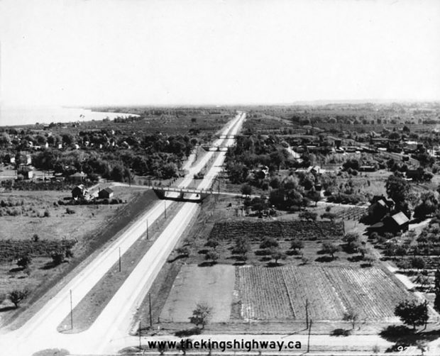 Photo noir et blanc de l’autoroute Queen Elizabeth Way, vers l’est ; zone de terres agricoles, avec le lac sur la gauche.