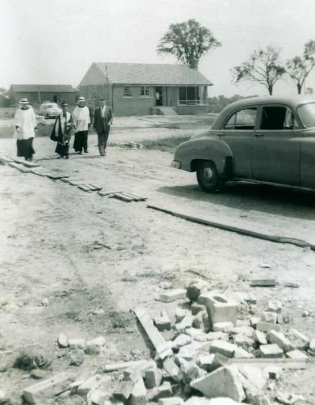 Photo noir et blanc de quatre hommes marchant vers une voiture ; trois prêtres et un homme en costume de ville.