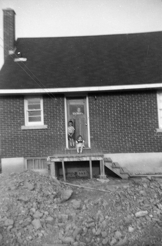 Photo noir et blanc de 2 enfants au dehors derrière la maison et d’une femme à la porte ; maison en cours de construction.