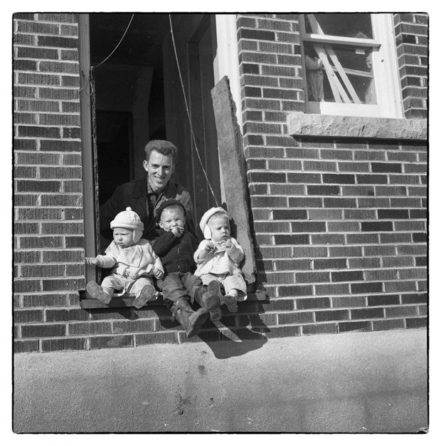 Photo noir et blanc d’un homme et de trois enfants assis près de la porte d’une maison en cours de construction.