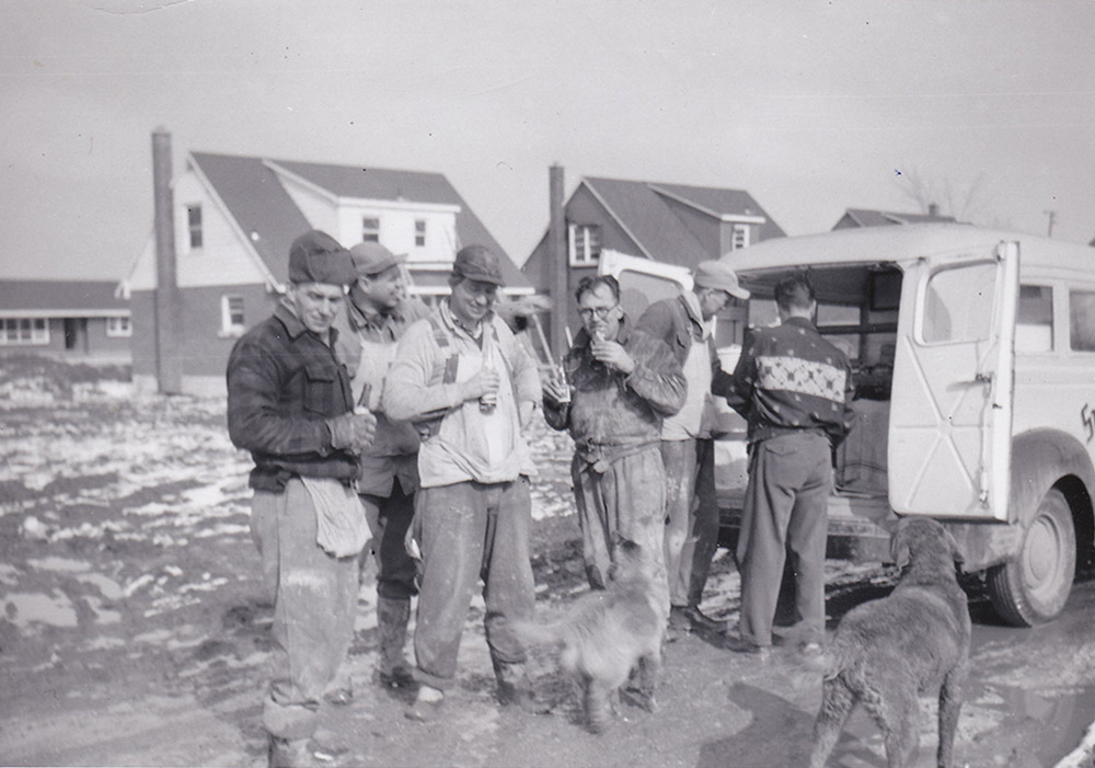 Photo noir et blanc d’un groupe d’hommes lors d’une pause sur un chantier de construction ; avec deux chiens au premier plan.