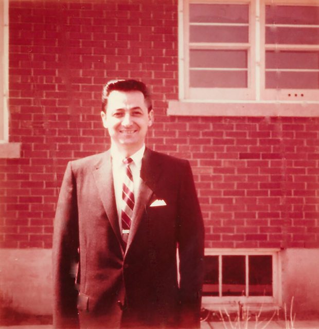 Photo couleur décolorée d’un homme en costume de ville devant une maison.