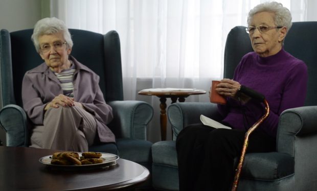 Photo couleur de deux femmes assises sur des fauteuils verts, se faisant face diagonalement, celle sur la droite avec une canne sur le bras et une tasse à la main.