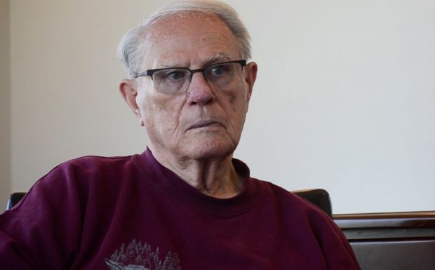 Photo couleur d’un homme portant des lunettes, assis et en chemise marron.