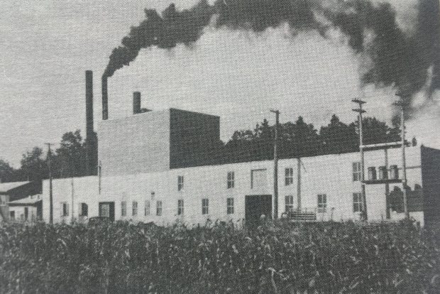 Photo en noir et blanc d'un long bâtiment d'usine muni de trois cheminées dont l'une rejette de la fumée. 