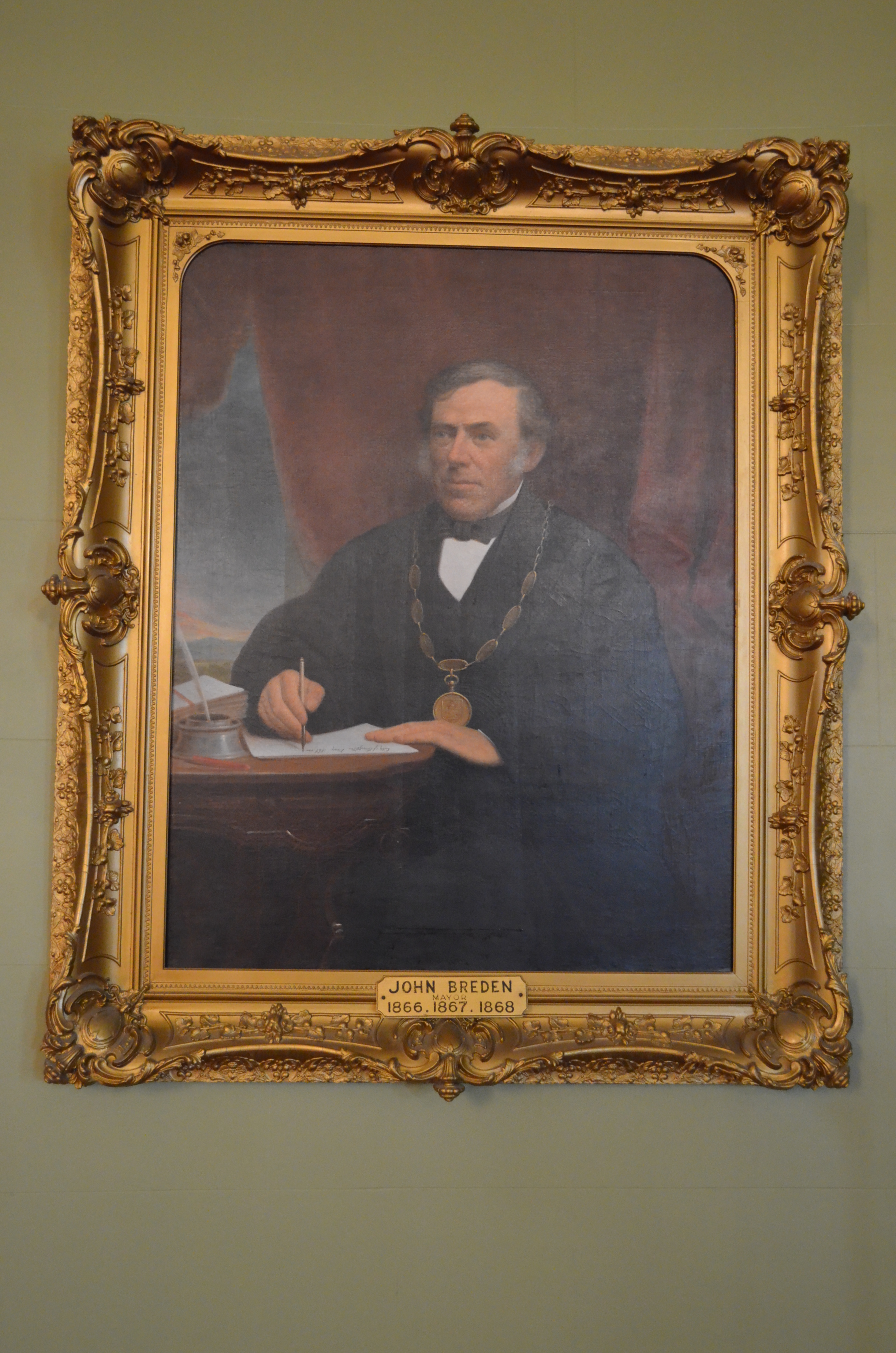 Portrait officiel de John Breden, maire de Kingston, assis à son bureau tenant une feuille et une plume.