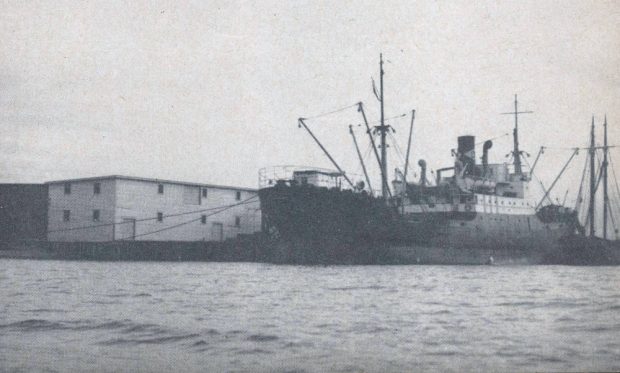 Photographie en noir et blanc d’un bateau à vapeur déchargeant du charbon, avec un entrepôt blanc à deux étages dans l’arrière-plan à gauche de la photo.