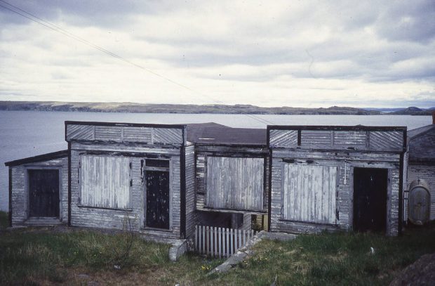 Photographie en couleur d’un magasin usé et délabré, les fenêtres couvertes de planches. L’océan et une côte lointaine forment l’arrière-plan de la photo.