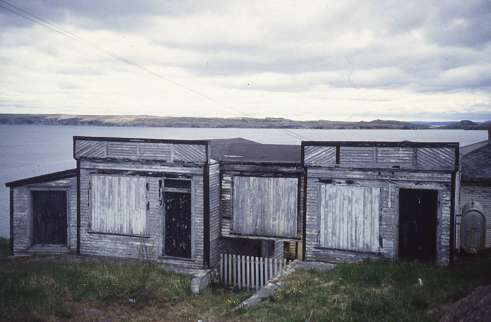 Photographie en couleur d’un magasin usé et délabré, les fenêtres couvertes de planches. L’océan et une côte lointaine forment l’arrière-plan de la photo.