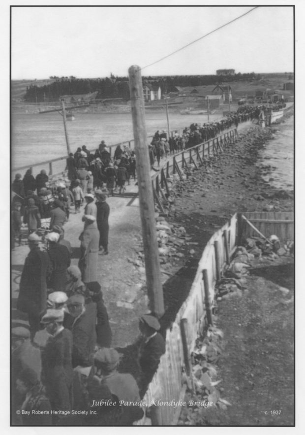 Photographie d’archive en noir et blanc d’une chaussée et d’un pont passant sur une étendue d’eau, avec une foule s’éloignant de l’objectif en paradant.