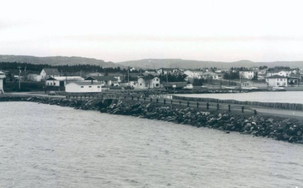 Photographie d’archive en noir et blanc d’une chaussée et d’un pont passant sur une étendue d’eau avec le rivage et des maisons en arrière-plan et des collines au loin.