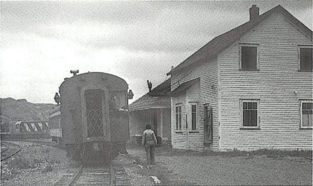 Photographie d’archive en noir et blanc montrant un wagon de train sur des rails à gauche, ainsi qu’un entrepôt à marchandises en bois d’un étage et un dépôt ferroviaire en bois de deux étages à droite.