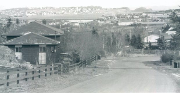 Photographie en noir et blanc montrant une route longée de clôtures. Il y a une maison à gauche de la photo et on voit une partie du village au loin, avec des collines et la mer à l’arrière.