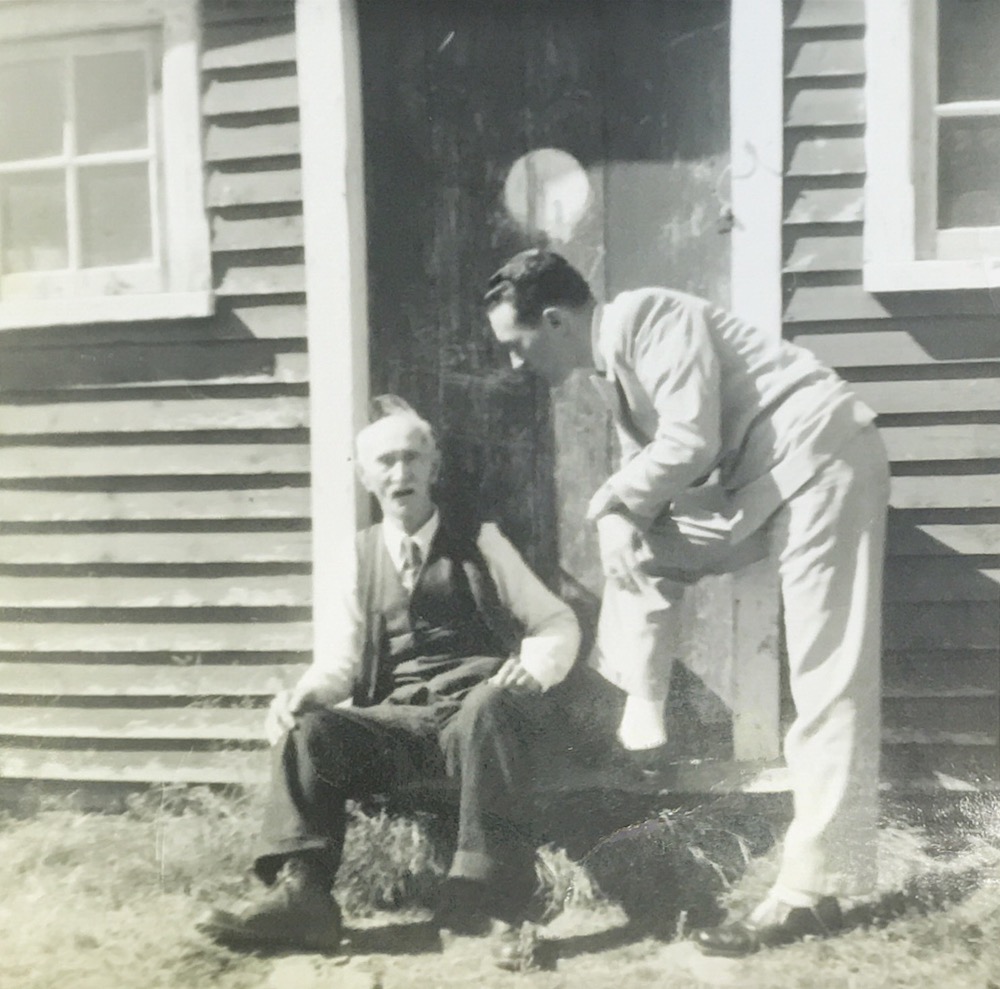 Photographie en noir et blanc de deux hommes se tenant sur le seuil de la forge Littlejohn. William H. Littlejohn est assis sur le seuil et son petit-fils, Bill Littlejohn, est debout à côté de lui, les mains posées sur un genou.