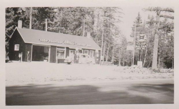 Photo en noir et blanc d’un bâtiment en rondins de bois avec des pompes à essence et une voiture ancienne devant; affiche à la droite; arbres à l'arrière-plan