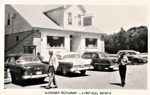 Photo en noir et blanc d’un édifice, avec des gens et des voitures anciennes à l'avant-plan et des arbres à l'arrière-plan