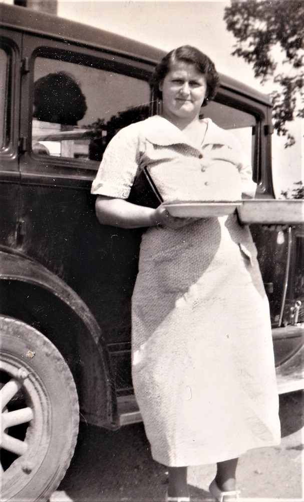 Photo en noir et blanc d’une femme en robe blanche qui tient deux tartes devant une voiture ancienne