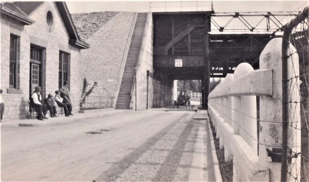 Photo en noir et blanc d’une voiture ancienne qui traverse le tunnel sous l'écluse-ascenseur, avec des bâtiments et des personnes à l’avant-plan