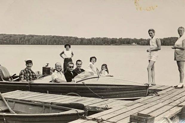 Photo en noir et blanc de touristes à bord d’un bateau à moteur en bois, lac à l’arrière-plan