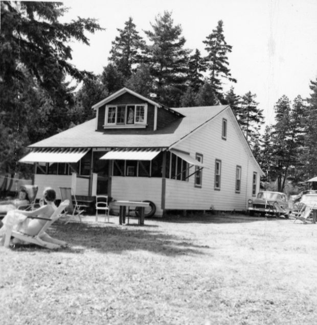 Photo en noir et blanc d’un bâtiment à charpente blanche, avec des gens qui sont assis sur des meubles de jardin à l'avant-plan et une voiture ancienne à l'arrière-plan