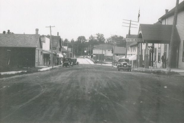 Photo en noir et blanc d’un paysage urbain, avec des voitures anciennes stationnées devant les édifices et des poteaux électriques bordant la rue