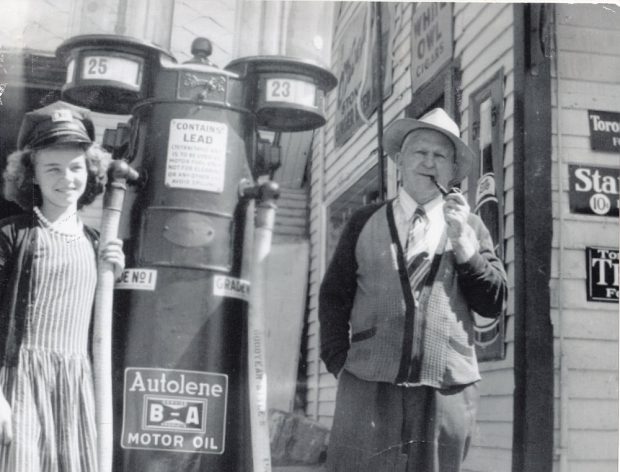 Photo en noir et blanc d’une jeune femme en uniforme et d’un homme plus âgé qui se tient debout devant une pompe à essence ancienne