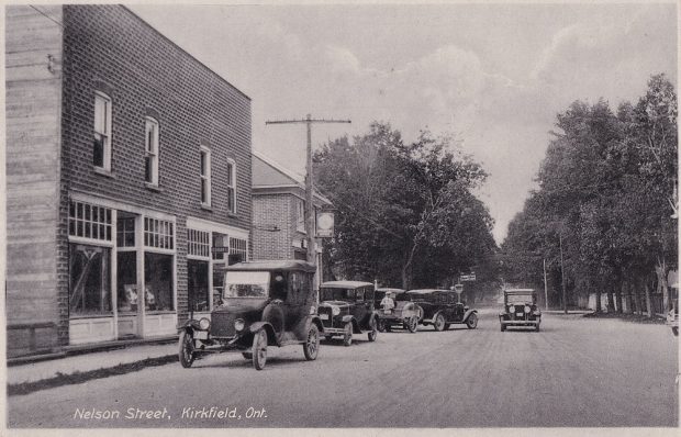 Photo en noir et blanc de voitures anciennes stationnées devant un édifice, avec des arbres à l’arrière-plan