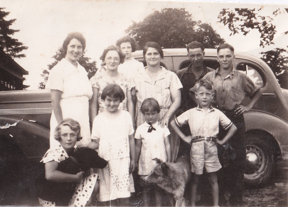 Photo en noir et blanc d’adultes, d’enfants et de deux chiens devant une voiture ancienne, avec des arbres à l'arrière-plan