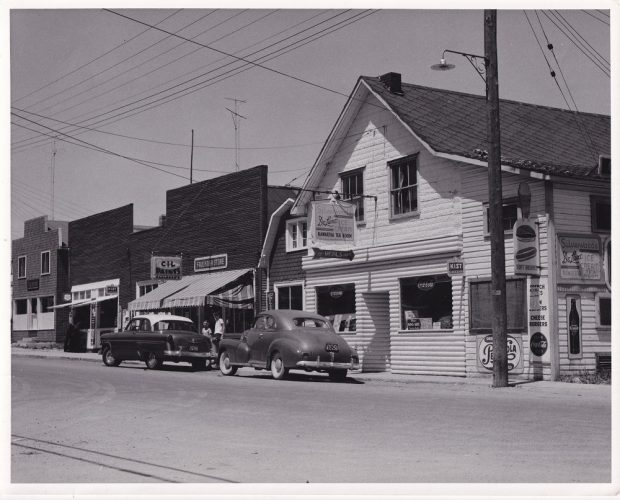 Photo en noir et blanc de bâtiments couverts d’affiches, avec deux voitures anciennes stationnées à l'avant-plan sous des fils électriques