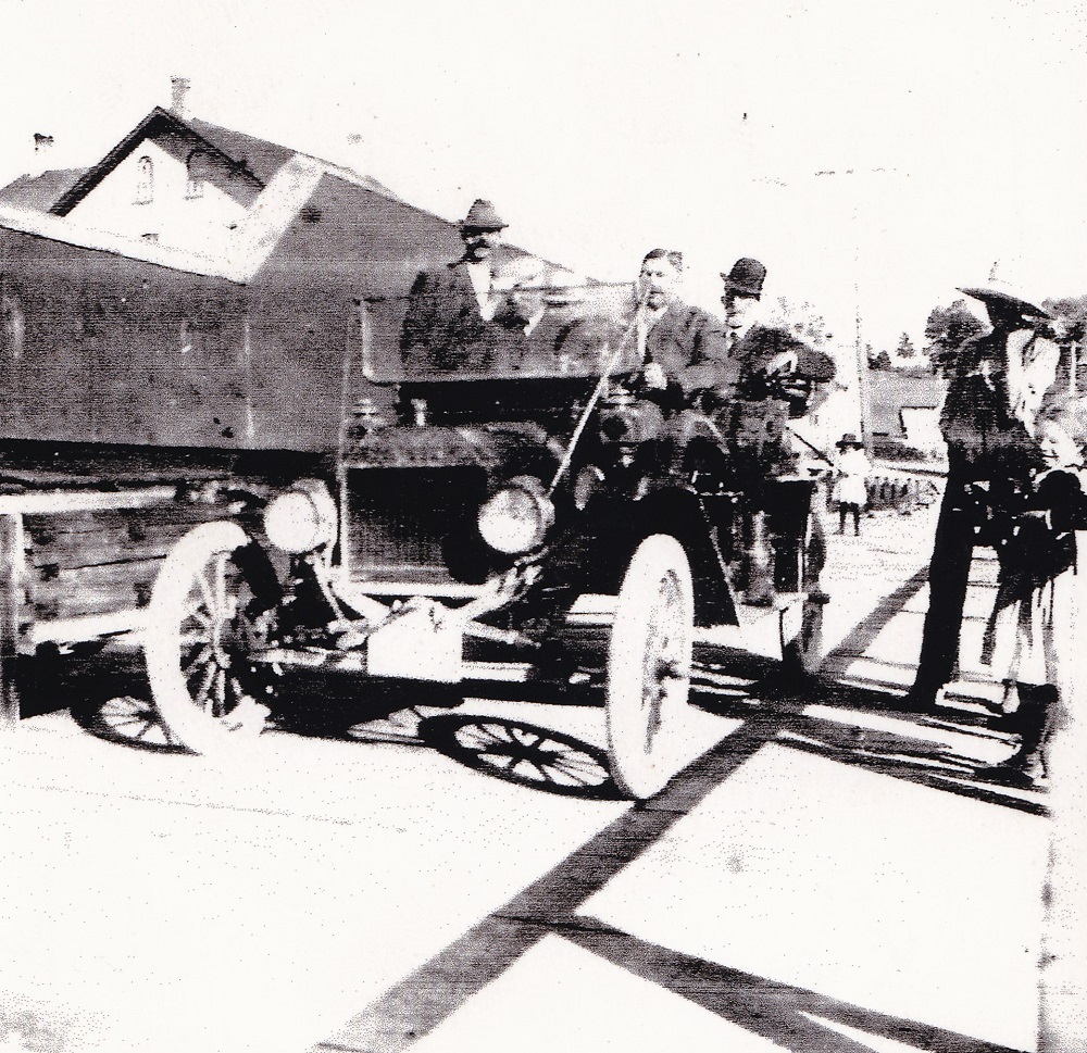 Photo en noir et blanc des passagers d’une voiture ancienne qui traverse un pont, avec un édifice à l’arrière-plan