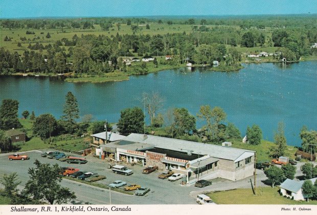 Photo aérienne en couleur qui montre le lac, un édifice et un parc de stationnement, avec des véhicules anciens